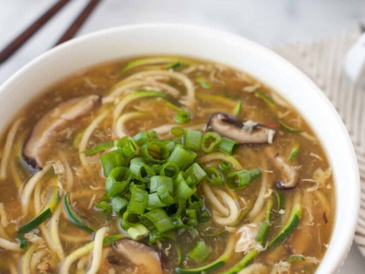 A bowl of noodle soup garnished with chopped green onions. The soup contains visible mushrooms and zucchini spirals in a light broth. A pair of chopsticks rests beside the bowl on a marble surface. Another bowl is partially visible in the background.