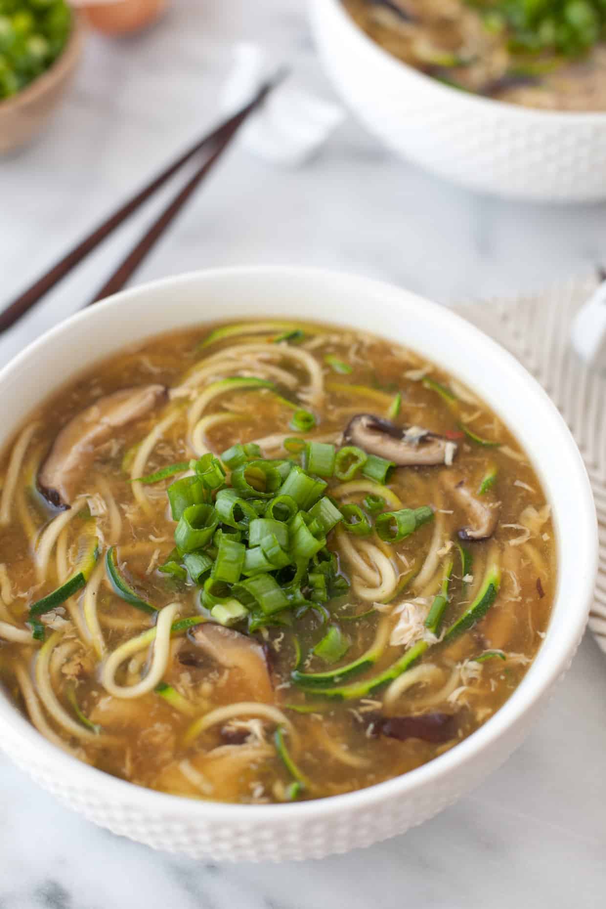 A bowl of noodle soup garnished with chopped green onions. The soup contains visible mushrooms and zucchini spirals in a light broth. A pair of chopsticks rests beside the bowl on a marble surface. Another bowl is partially visible in the background.