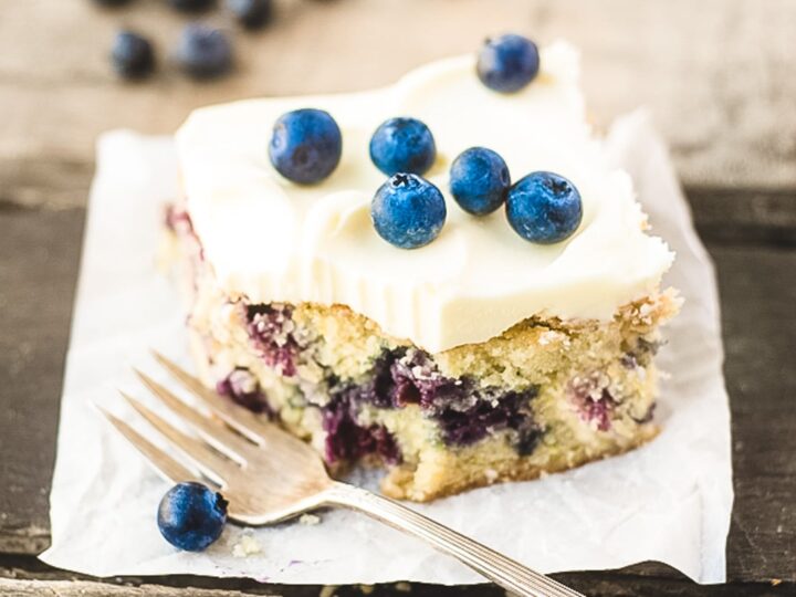 A slice of blueberry cake with creamy frosting topped with fresh blueberries on a piece of parchment paper. A fork rests beside it. Additional blueberries are scattered in the background on a wooden surface.