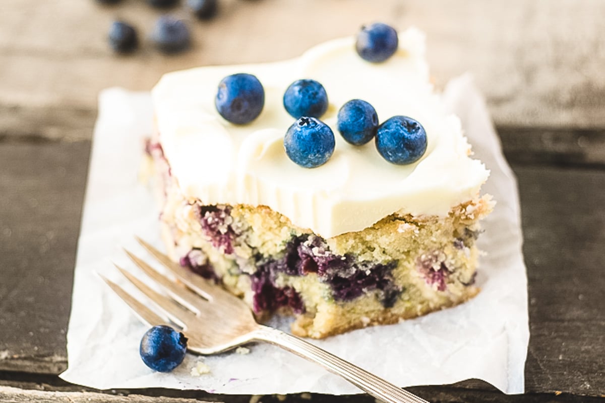A slice of blueberry cake with creamy frosting topped with fresh blueberries on a piece of parchment paper. A fork rests beside it. Additional blueberries are scattered in the background on a wooden surface.