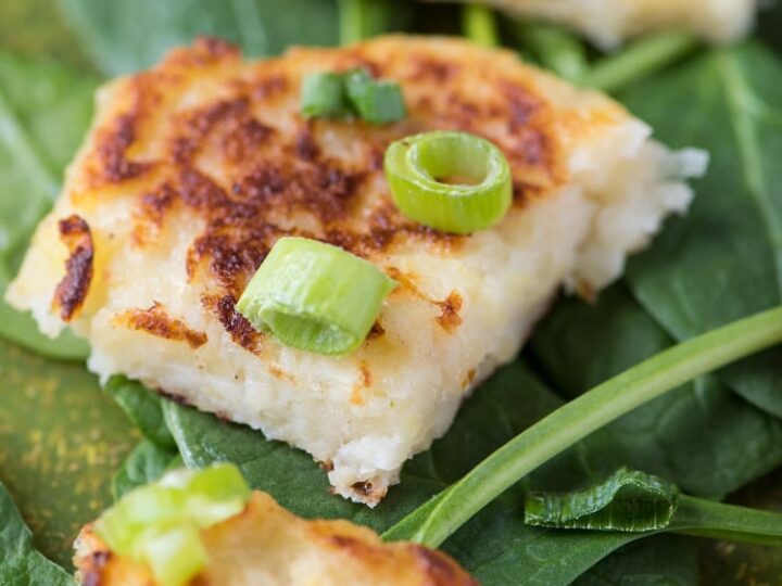 Close-up of crispy potato pancakes topped with chopped green onions, placed on a bed of fresh spinach leaves. The pancakes are golden brown with a soft, fluffy interior visible.