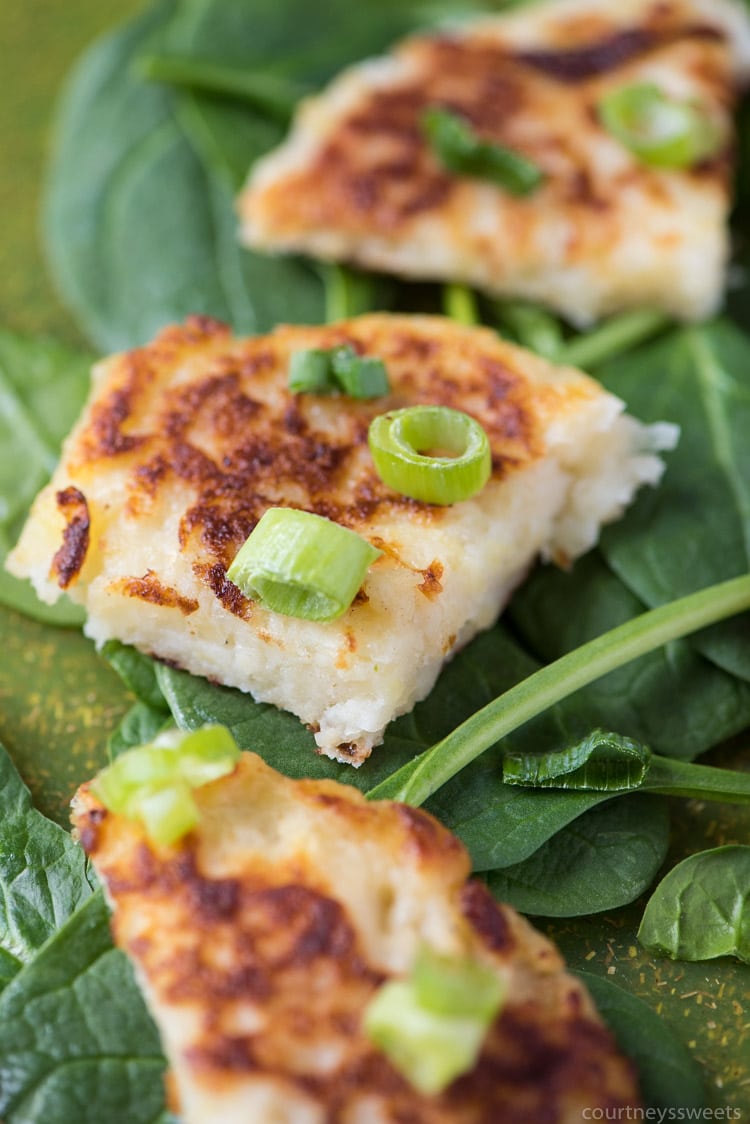 Close-up of crispy potato pancakes topped with chopped green onions, placed on a bed of fresh spinach leaves. The pancakes are golden brown with a soft, fluffy interior visible.