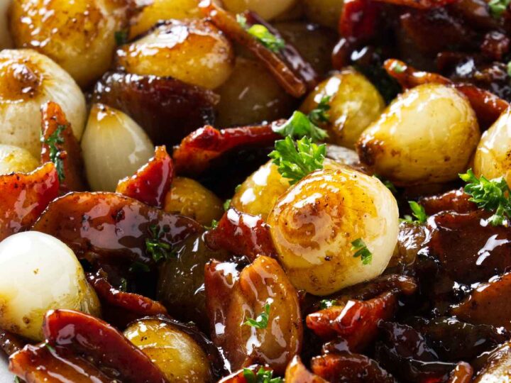 A bowl of glazed onions and crispy bacon pieces garnished with fresh herbs. The onions have a shiny, caramelized appearance, and the bacon is browned. The dish is served in a white bowl on a light surface.