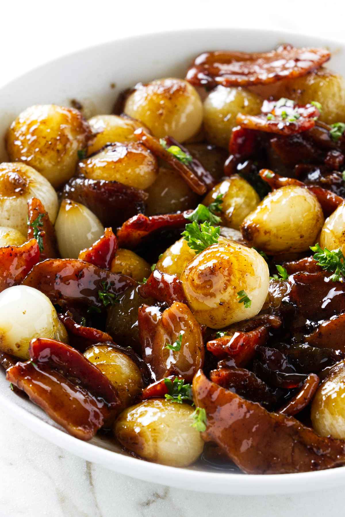 A bowl of glazed onions and crispy bacon pieces garnished with fresh herbs. The onions have a shiny, caramelized appearance, and the bacon is browned. The dish is served in a white bowl on a light surface.