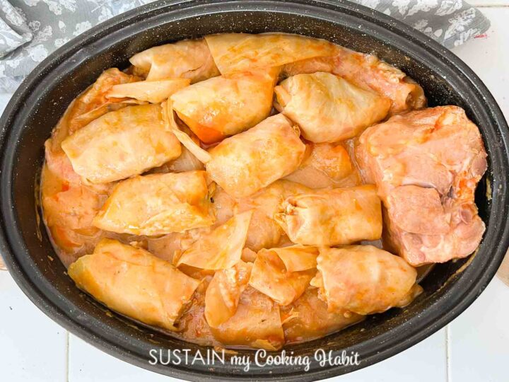 A pot filled with cooked cabbage rolls in tomato sauce. The rolls are tightly packed and have a glossy appearance, with some visible orange-hued sauce. The pot is placed on a light-colored surface.