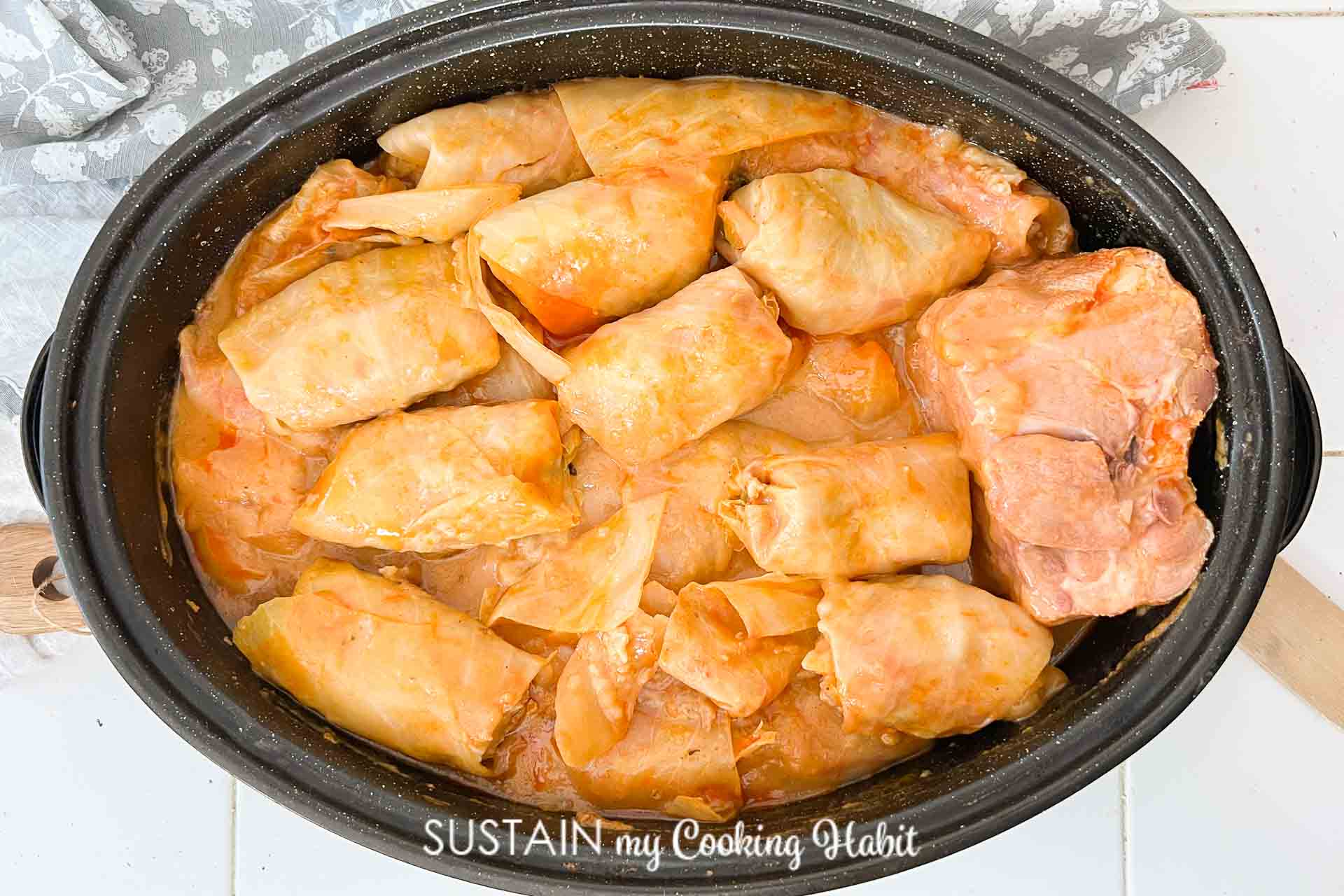 A pot filled with cooked cabbage rolls in tomato sauce. The rolls are tightly packed and have a glossy appearance, with some visible orange-hued sauce. The pot is placed on a light-colored surface.
