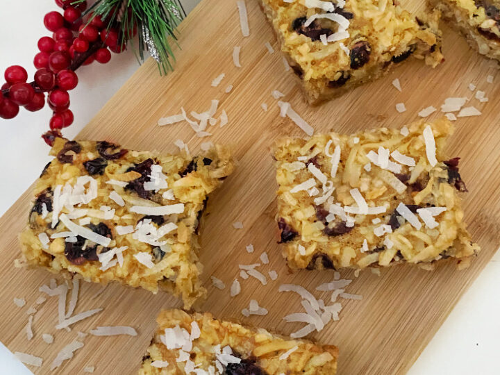 Festive dessert bars topped with shredded coconut and cranberries are placed on a wooden board. A decorative pine and berry arrangement sits to the side, adding a holiday touch.