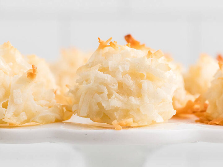 A close-up of several coconut macaroons resting on a white plate. The macaroons are golden brown on top, indicating they are freshly baked. The background is blurred, focusing attention on the texture of the macaroons.