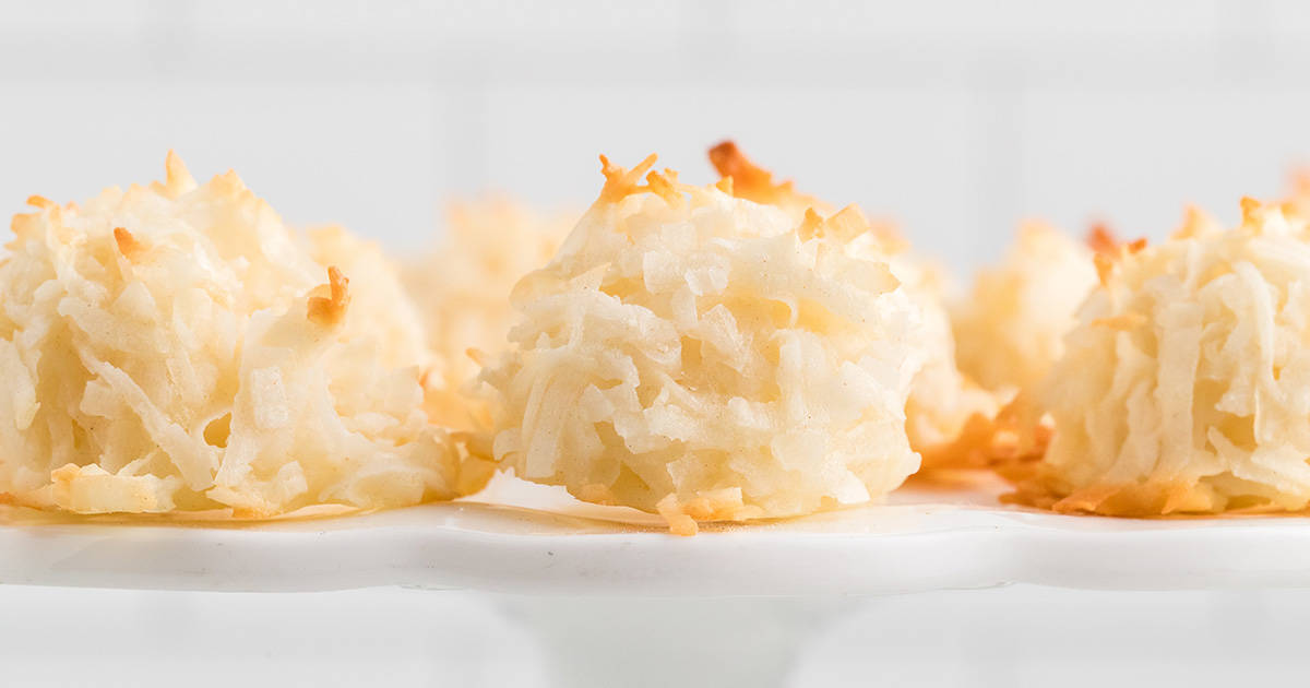 A close-up of several coconut macaroons resting on a white plate. The macaroons are golden brown on top, indicating they are freshly baked. The background is blurred, focusing attention on the texture of the macaroons.