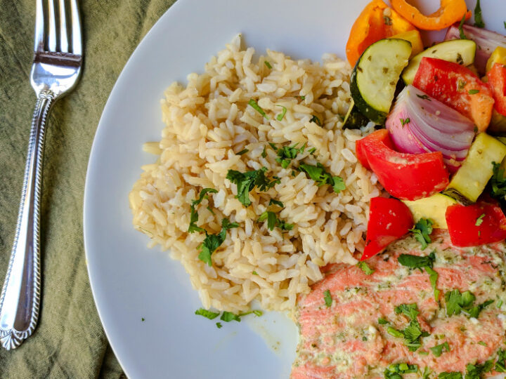 A plate featuring a serving of brown rice garnished with herbs, colorful roasted vegetables including zucchini, red onion, and bell peppers, alongside a portion of seasoned salmon. A fork is placed on a green cloth napkin beside the plate.