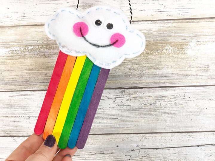 A hand holding a homemade craft featuring a smiling cloud made of white felt with black eyes and pink cheeks. Rainbow-colored popsicle sticks hang from the cloud. The background is a light wooden surface.