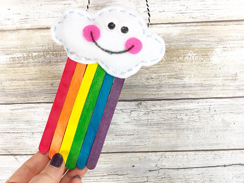 A hand holding a homemade craft featuring a smiling cloud made of white felt with black eyes and pink cheeks. Rainbow-colored popsicle sticks hang from the cloud. The background is a light wooden surface.