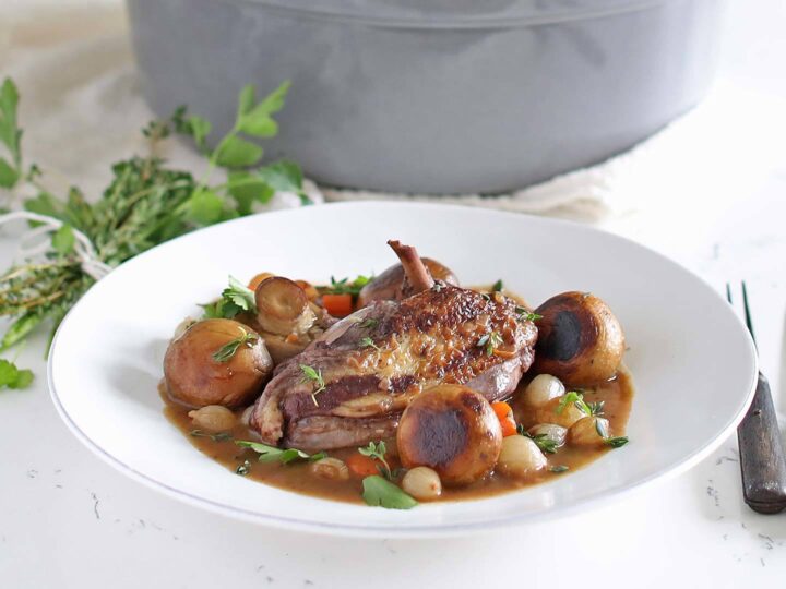 A white plate with a serving of coq au vin, featuring a cooked chicken piece, mushrooms, pearl onions, and carrots garnished with fresh herbs. A large pot is in the background with cutlery placed beside the plate.