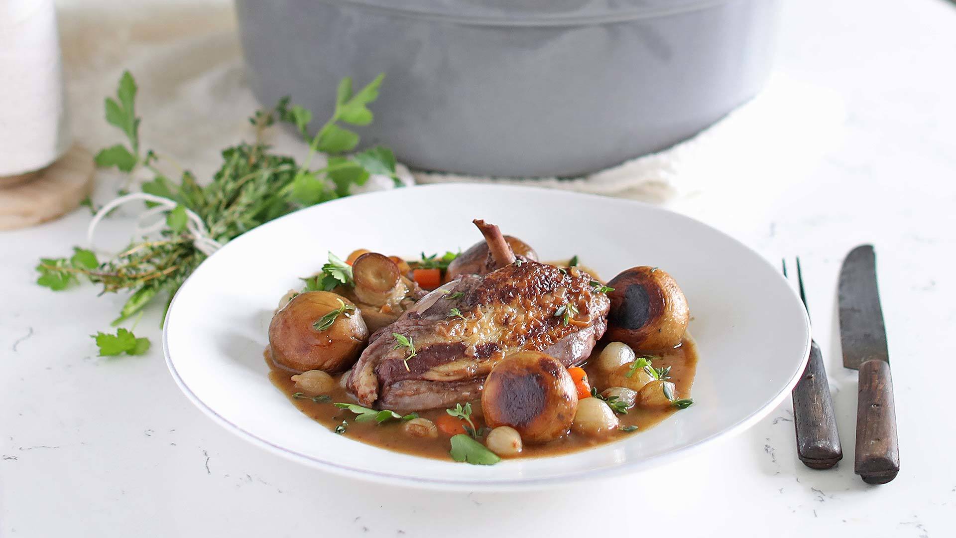 A white plate with a serving of coq au vin, featuring a cooked chicken piece, mushrooms, pearl onions, and carrots garnished with fresh herbs. A large pot is in the background with cutlery placed beside the plate.