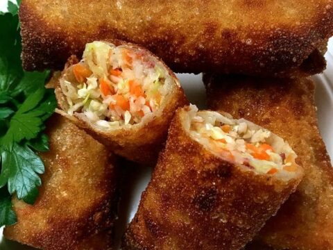 Crispy golden-brown egg rolls are stacked on a white plate. One is cut open, revealing a filling of shredded cabbage, carrots, and other vegetables. Fresh parsley garnishes the plate.