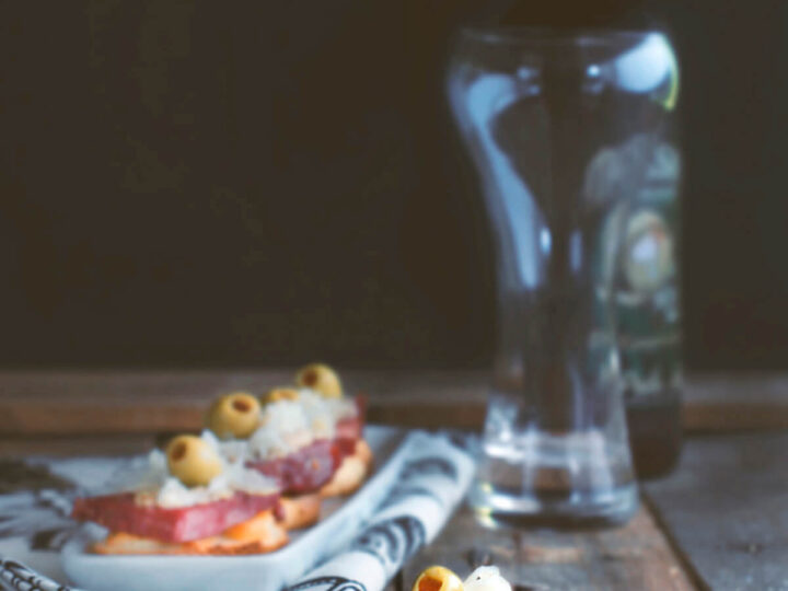 A rustic setting with a serving of sliced cured meats, cheese, and green olives on a napkin placed on a wooden surface. A bottle of dark beer stands in the background, next to an empty glass. A small knife is on the table.