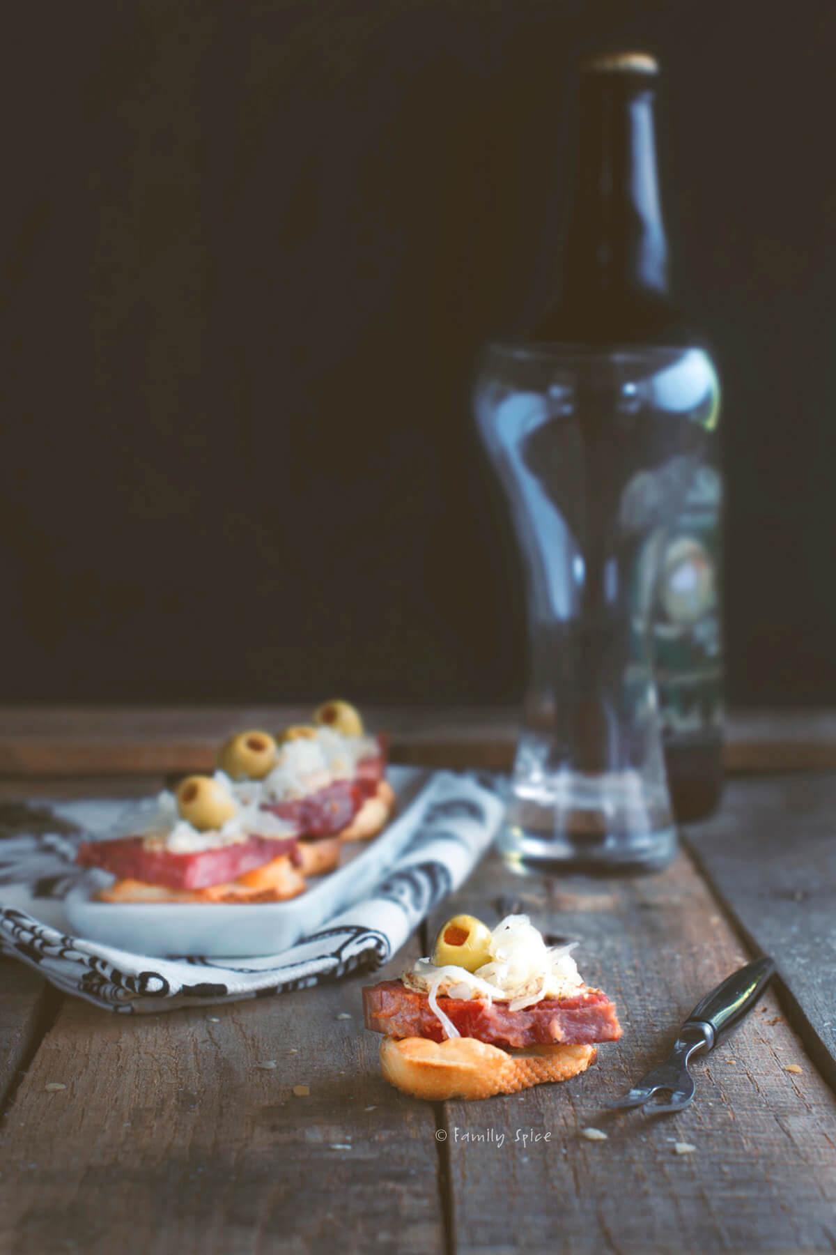 A rustic setting with a serving of sliced cured meats, cheese, and green olives on a napkin placed on a wooden surface. A bottle of dark beer stands in the background, next to an empty glass. A small knife is on the table.