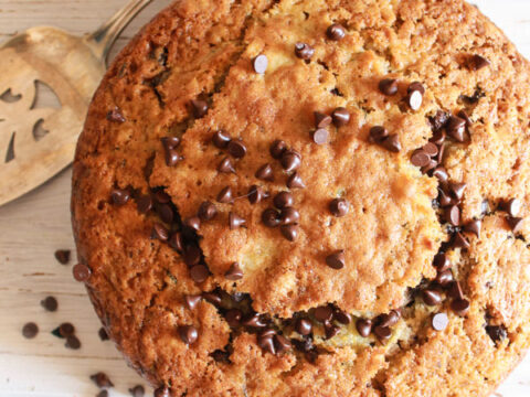 Overhead view of a round chocolate chip cake on a light wooden surface. The cake is topped with scattered chocolate chips. A silver serving spatula is partially visible beside the cake.