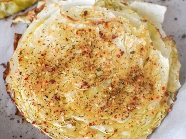 A close-up of a roasted cabbage slice on a baking sheet. The cabbage is seasoned with herbs and spices, giving it a golden-brown color with visible flakes of seasoning. Edges are slightly crispy.