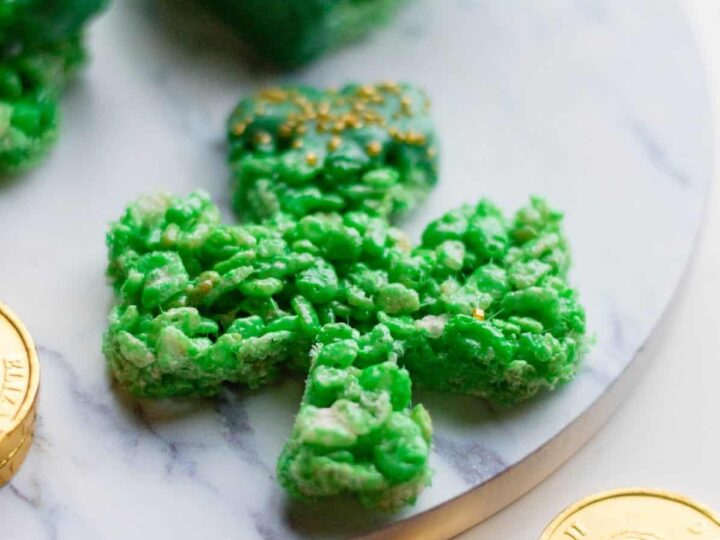 Shamrock-shaped green rice cereal treat on a marble platter, surrounded by chocolate gold coins. Additional treats stacked in the background.