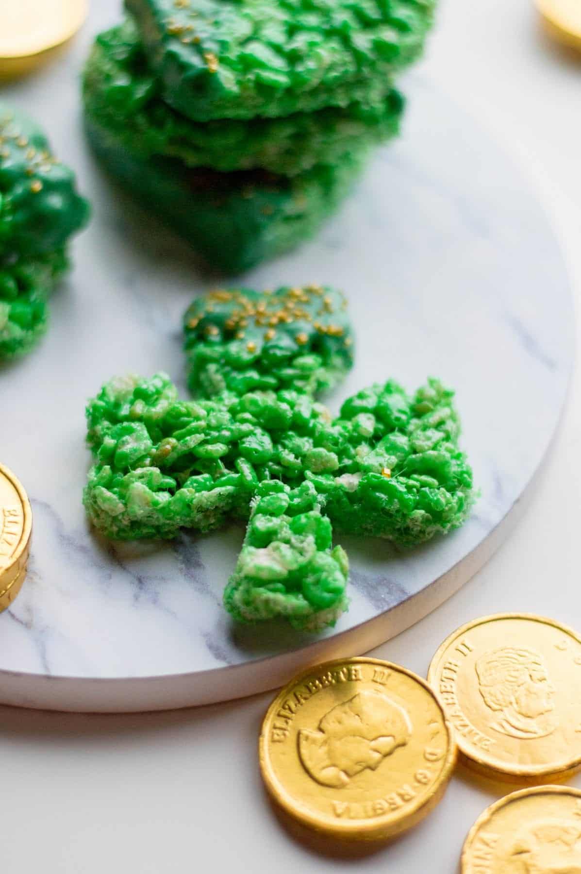 Shamrock-shaped green rice cereal treat on a marble platter, surrounded by chocolate gold coins. Additional treats stacked in the background.