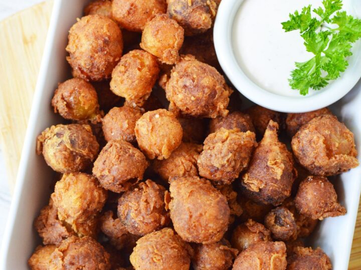 A rectangular white dish filled with golden-brown, round fried hushpuppies. A small bowl of creamy dipping sauce garnished with parsley is placed on the side. The dish is set on a wooden board.