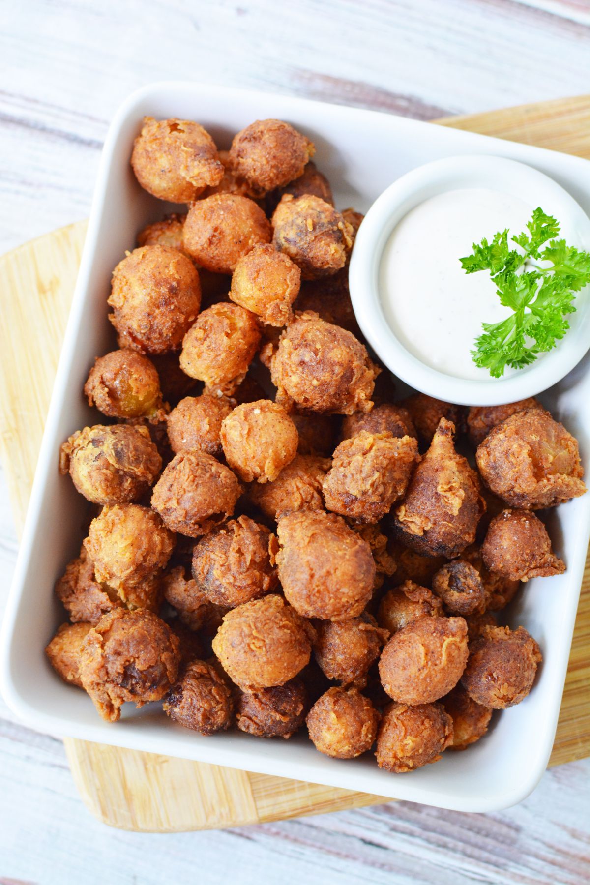 A rectangular white dish filled with golden-brown, round fried hushpuppies. A small bowl of creamy dipping sauce garnished with parsley is placed on the side. The dish is set on a wooden board.