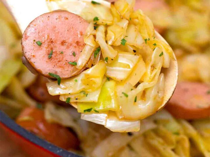 A wooden spoon holds a serving of cooked cabbage and sliced sausage garnished with herbs. In the background, a skillet filled with the same dish is partially visible.