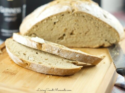 A loaf of sliced bread on a wooden cutting board, with two slices in the foreground. A knife is placed beside the bread. A jar is blurred in the background.
