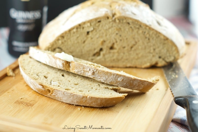 A loaf of sliced bread on a wooden cutting board, with two slices in the foreground. A knife is placed beside the bread. A jar is blurred in the background.