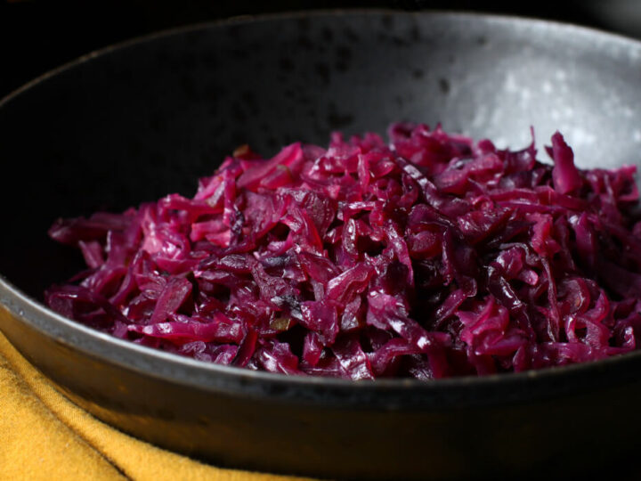 A black bowl filled with cooked red cabbage, placed on a mustard-colored cloth. The cabbage appears glossy and is finely shredded, indicating it is likely seasoned or sautéed. The background is dark, emphasizing the vibrant purple of the cabbage.