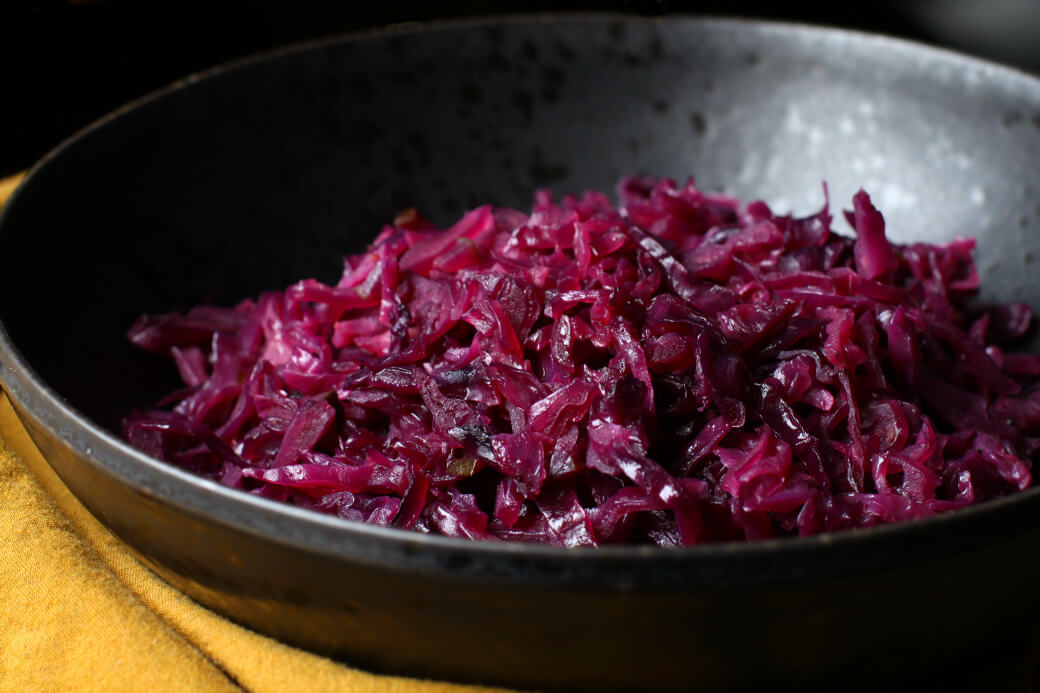 A black bowl filled with cooked red cabbage, placed on a mustard-colored cloth. The cabbage appears glossy and is finely shredded, indicating it is likely seasoned or sautéed. The background is dark, emphasizing the vibrant purple of the cabbage.