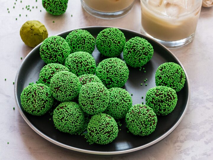 A black plate filled with green, sprinkle-coated truffles on a white surface. Two glasses of iced milk are in the background. A few truffles and sprinkles are scattered around the plate.