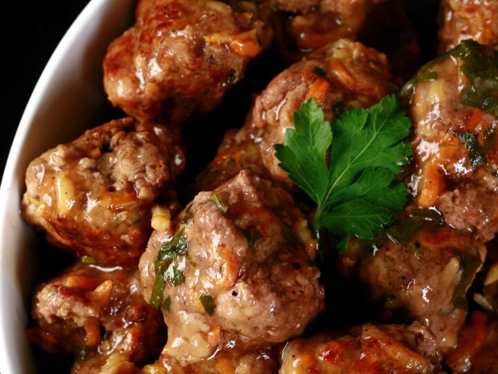 Close-up of a bowl filled with glazed meatballs garnished with a sprig of parsley. The meatballs are coated in a savory sauce, giving them a shiny appearance.