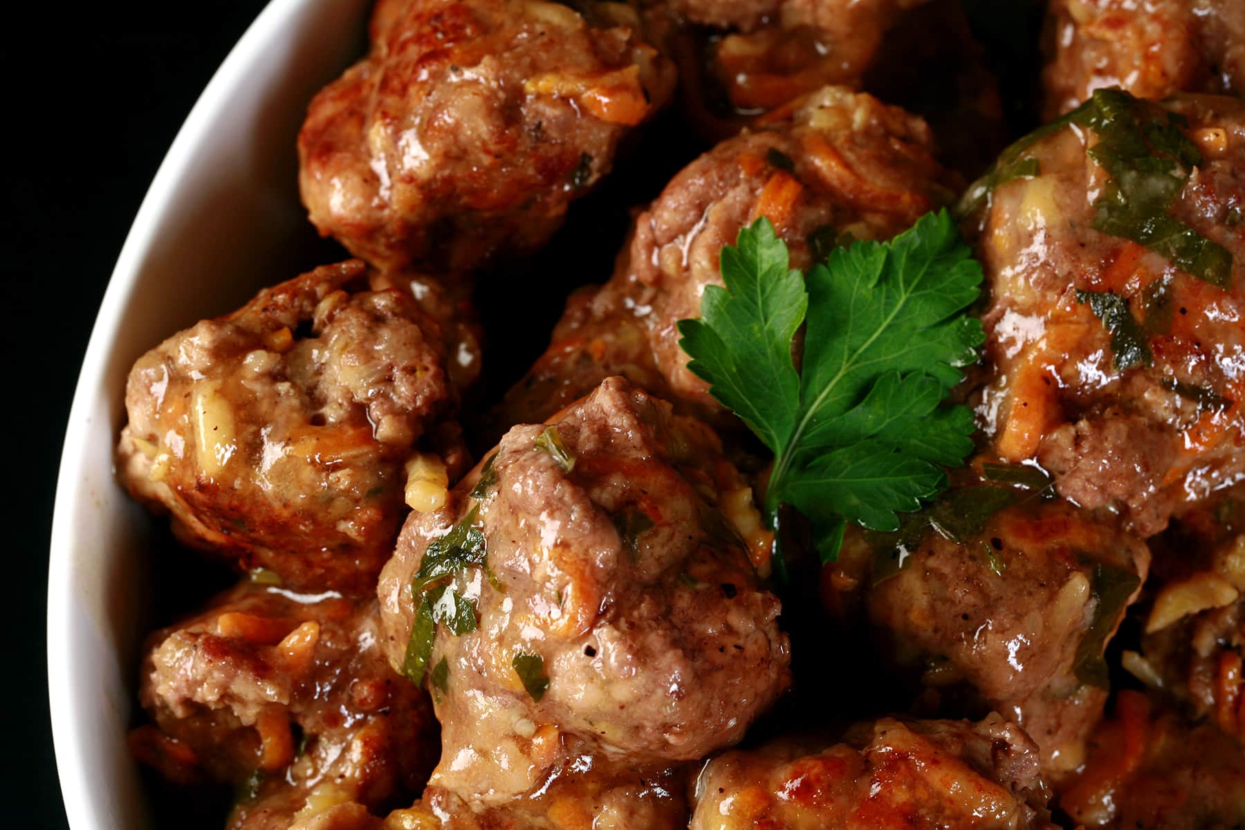 Close-up of a bowl filled with glazed meatballs garnished with a sprig of parsley. The meatballs are coated in a savory sauce, giving them a shiny appearance.