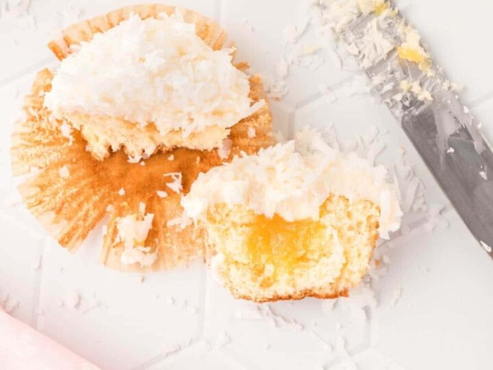 A partially eaten cupcake with coconut frosting is placed on a partially opened paper liner. There is a filling visible inside the cupcake. Shredded coconut is scattered around, and a knife with frosting rests beside it on a white surface.