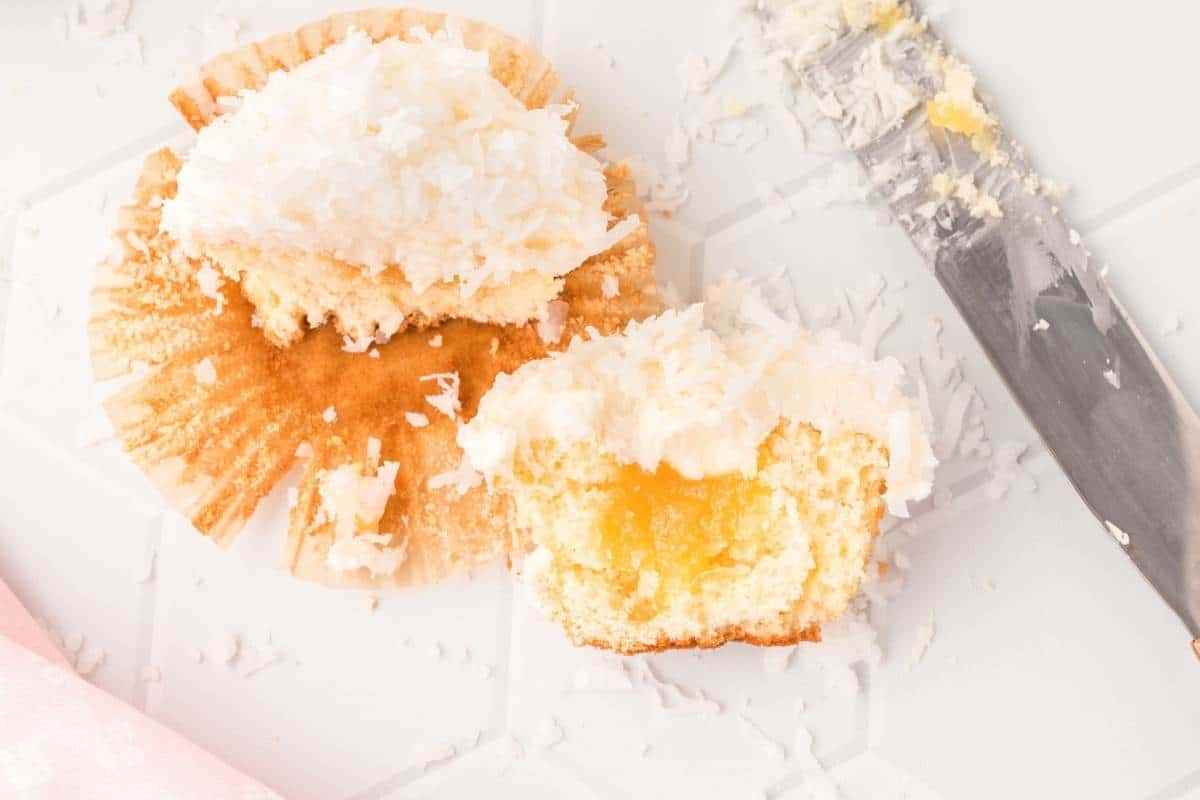 A partially eaten cupcake with coconut frosting is placed on a partially opened paper liner. There is a filling visible inside the cupcake. Shredded coconut is scattered around, and a knife with frosting rests beside it on a white surface.