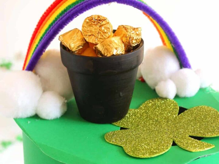 A small black pot filled with gold-wrapped chocolates sits on a green platform adorned with a gold glitter shamrock. A rainbow made of colorful pipe cleaners arches over the pot, and white cotton balls resemble clouds.
