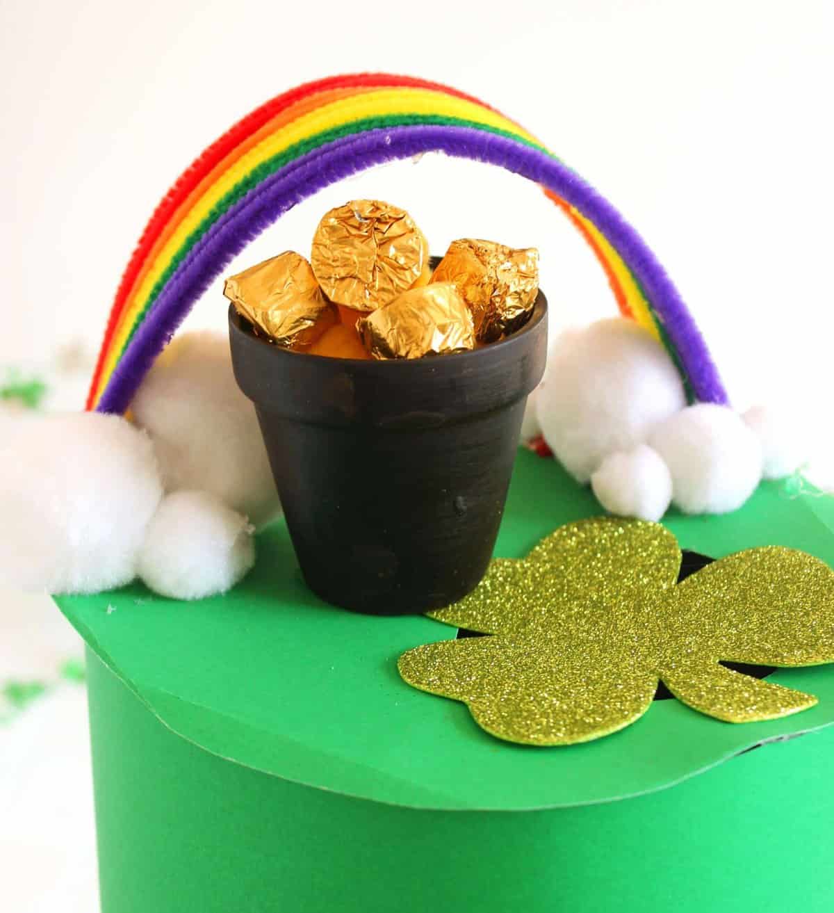 A small black pot filled with gold-wrapped chocolates sits on a green platform adorned with a gold glitter shamrock. A rainbow made of colorful pipe cleaners arches over the pot, and white cotton balls resemble clouds.