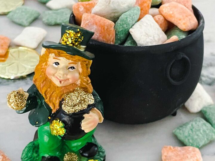A small leprechaun figurine with a green outfit and hat stands next to a black pot filled with gold coins and pastel-colored candies. More candies and coins are scattered on the marble surface around them.