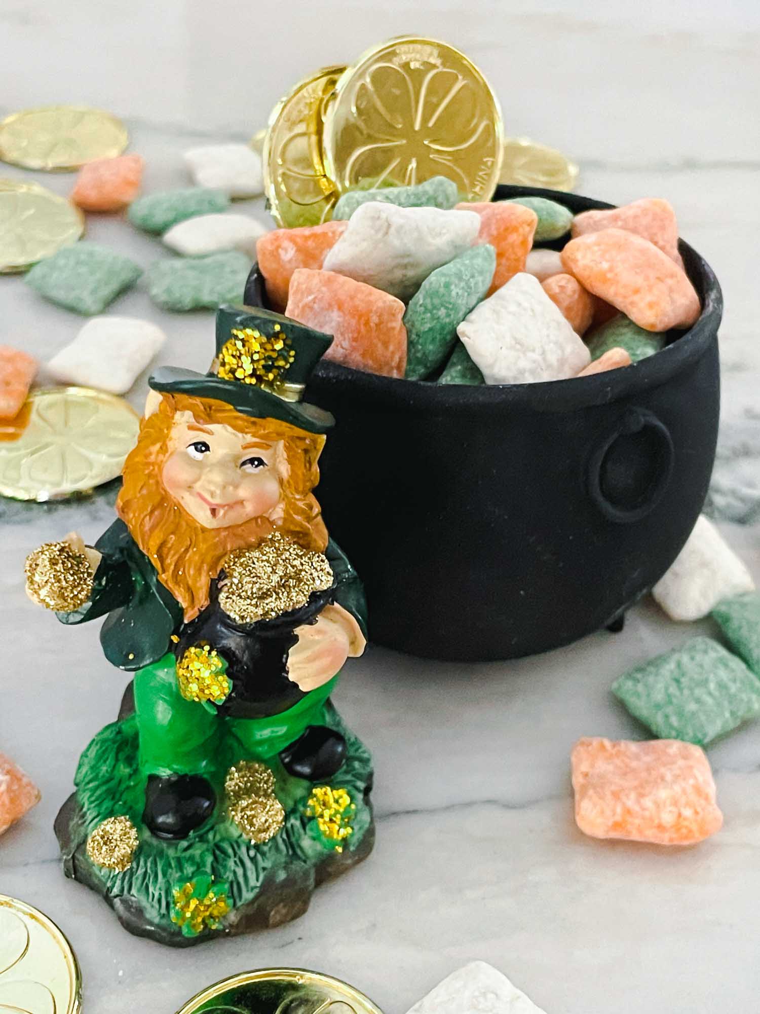A small leprechaun figurine with a green outfit and hat stands next to a black pot filled with gold coins and pastel-colored candies. More candies and coins are scattered on the marble surface around them.