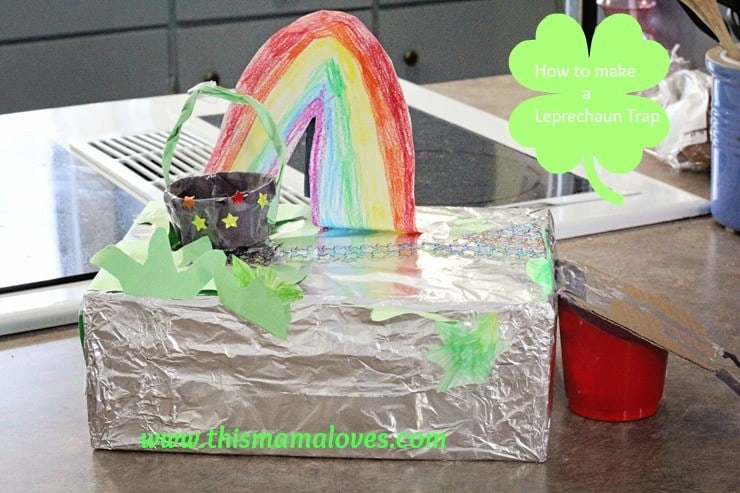 A homemade leprechaun trap made from a foil-covered box. It features a colorful rainbow leading to a black pot with stars, surrounded by green shamrocks. A small sign reads, How to make a Leprechaun Trap.