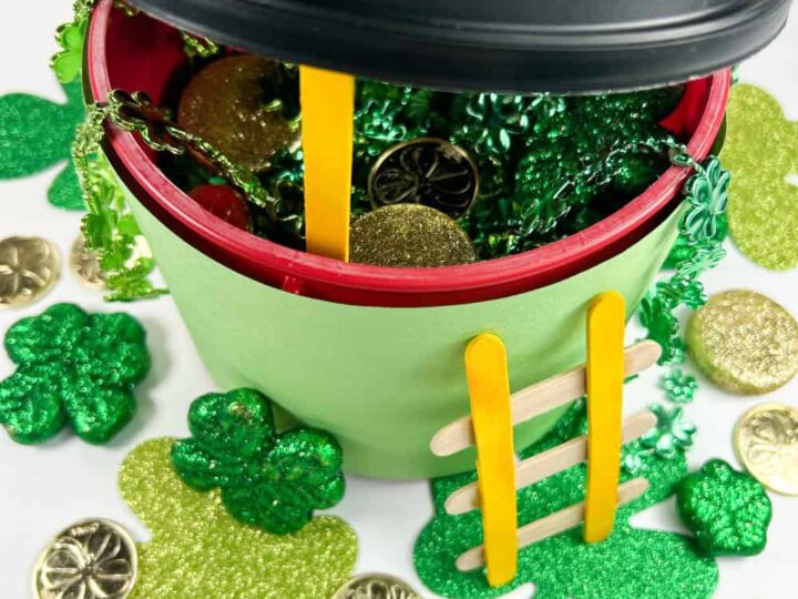 A festive pot decorated with green paper and yellow popsicle stick ladder, filled with green and gold coins. Surrounding the pot are green shamrocks and gold coins, creating a St. Patricks Day theme.