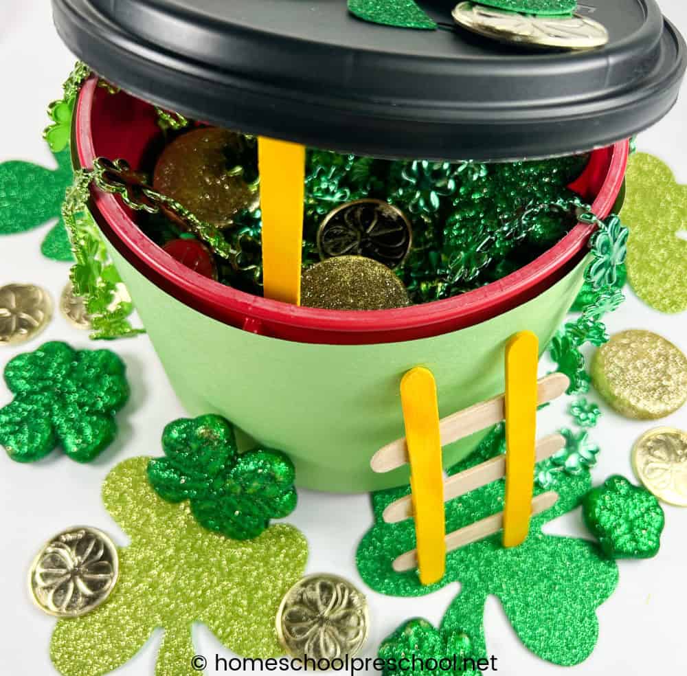 A festive pot decorated with green paper and yellow popsicle stick ladder, filled with green and gold coins. Surrounding the pot are green shamrocks and gold coins, creating a St. Patricks Day theme.