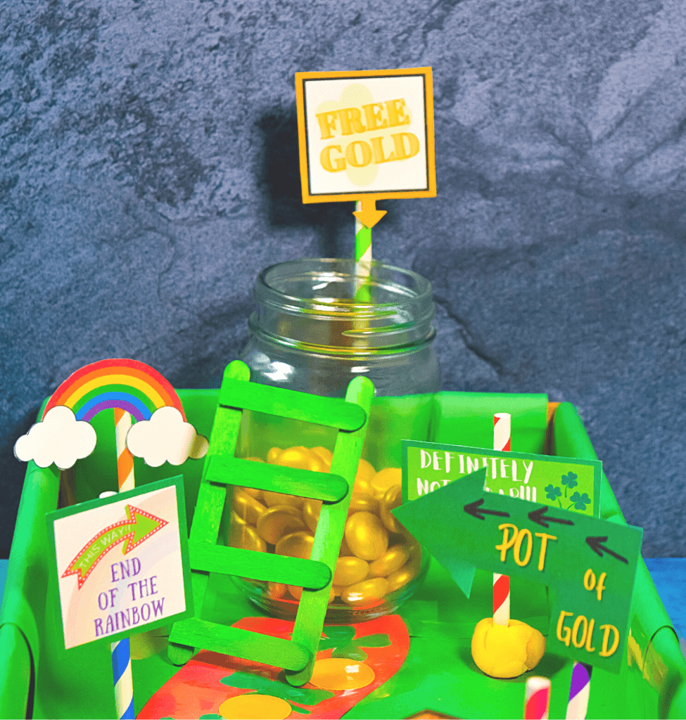 A playful setup with a jar of gold candies labeled Free Gold on a rainbow-colored board, perfect for leprechaun trap ideas. Signs read End of the Rainbow and Pot of Gold, while a small green ladder tempting any wandering leprechaun leans against the jar.