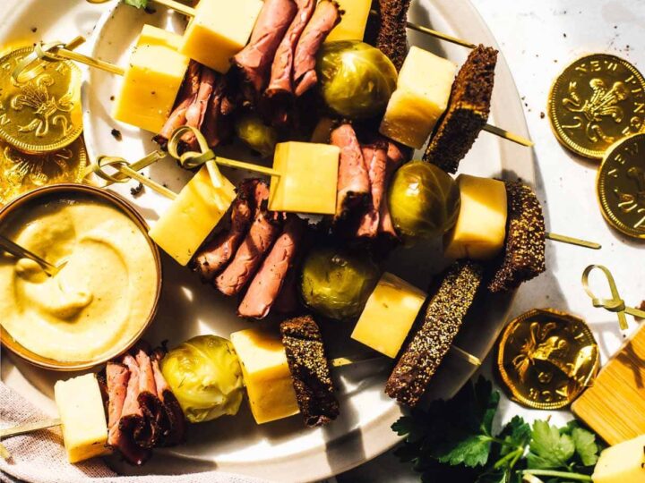 A plate with skewers featuring cheese cubes, rolled meat slices, olives, and brown bread. A small bowl of mustard is beside the skewers. Chocolate gold coins and fresh parsley are scattered around, with a textured cloth partially visible.