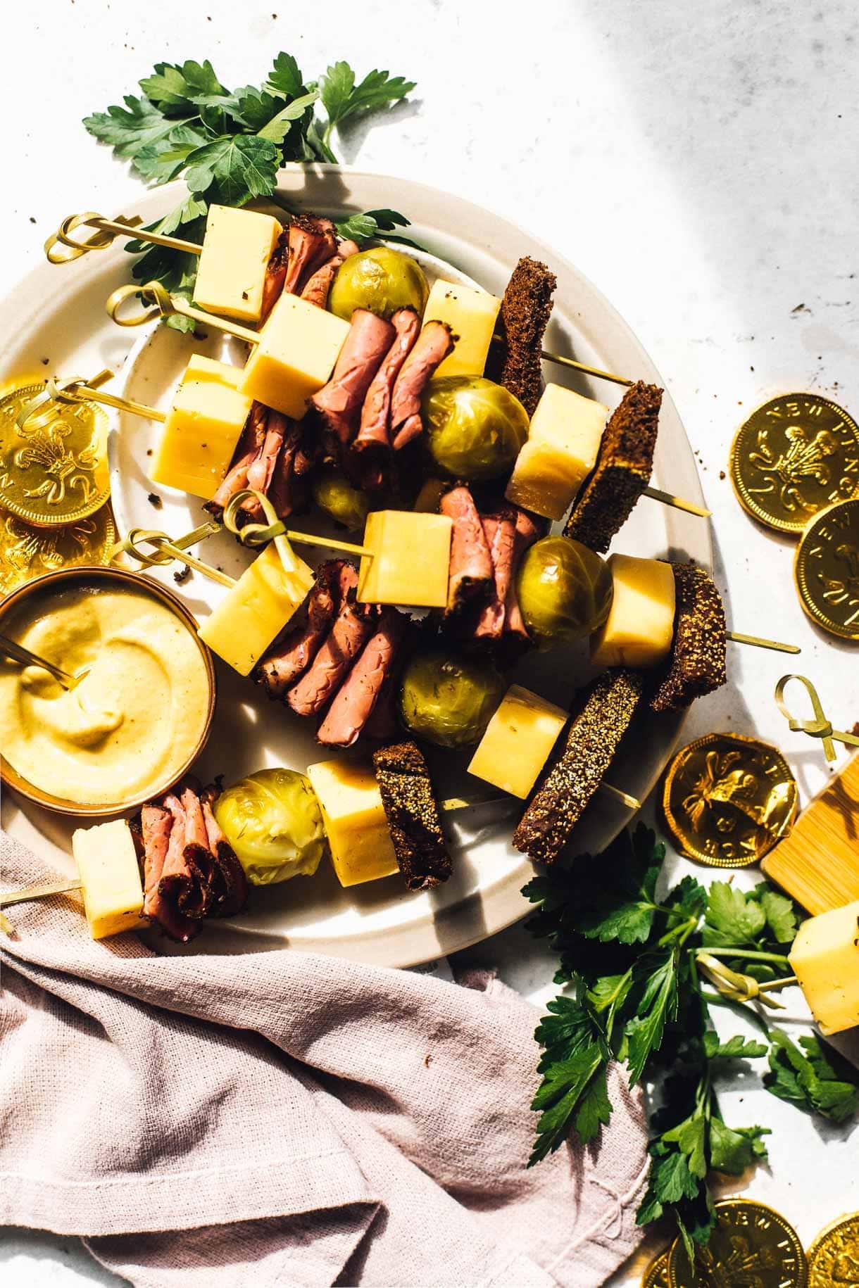 A plate with skewers featuring cheese cubes, rolled meat slices, olives, and brown bread. A small bowl of mustard is beside the skewers. Chocolate gold coins and fresh parsley are scattered around, with a textured cloth partially visible.