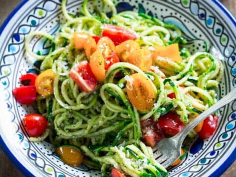A colorful bowl of spiralized zucchini noodles mixed with cherry tomatoes in shades of red and yellow, garnished with pesto sauce. The dish is served in a decorative ceramic bowl with a fork on the side.