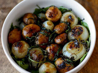 A white bowl filled with glazed caramelized pearl onions and leafy greens. The onions are golden brown with a shiny finish, suggesting they are cooked in a sauce. The bowl is placed on a wooden surface.