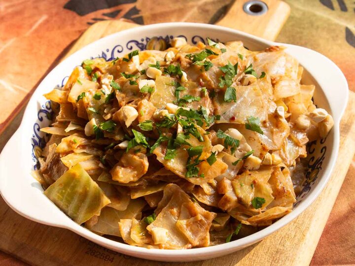 A white bowl filled with stir-fried cabbage and diced chicken, topped with chopped peanuts and fresh parsley. The dish is placed on a wooden board with a colorful tablecloth in the background.